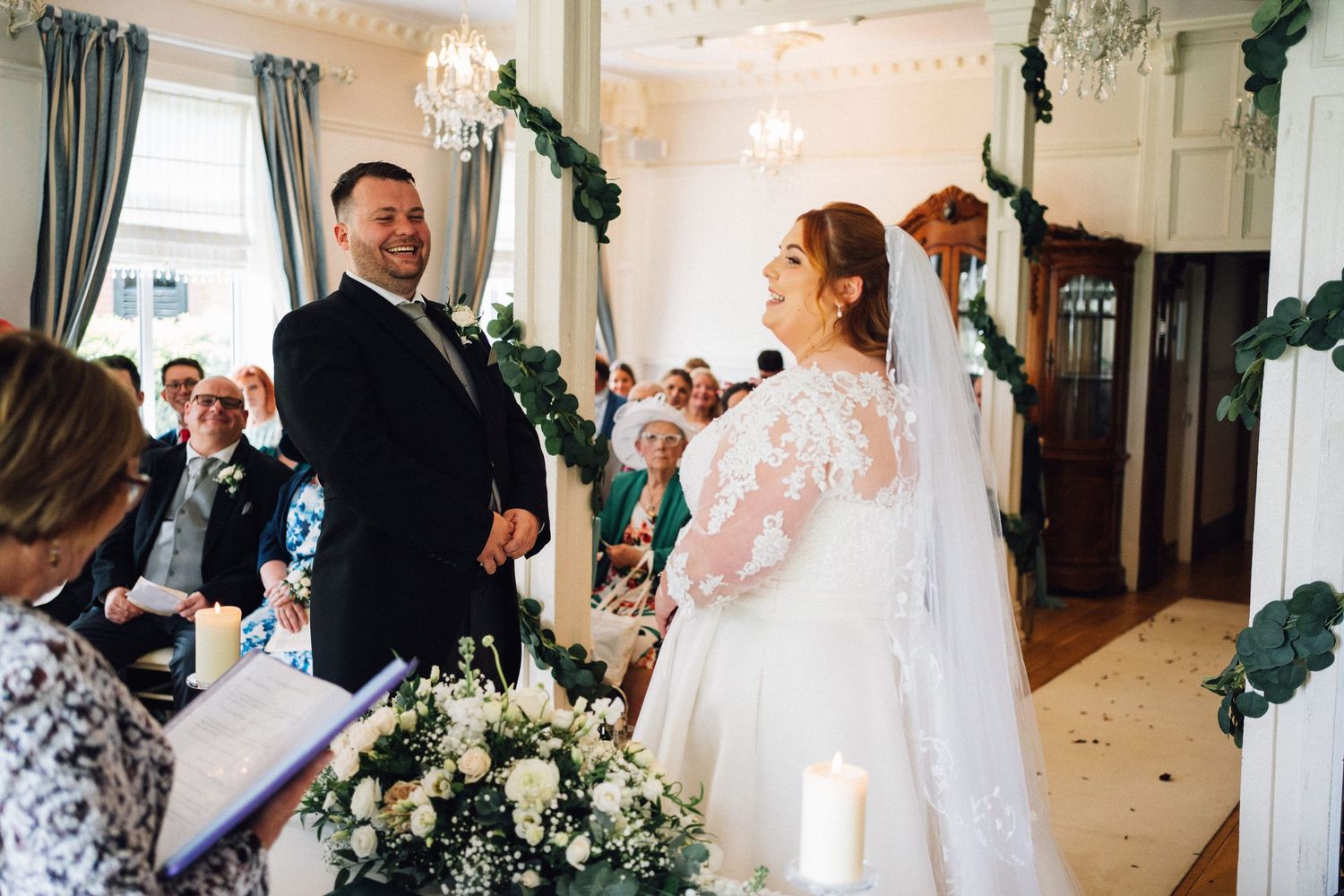 Bride and groom exchanging vows at elegant wedding ceremony with floral arrangements and guests seated in background.