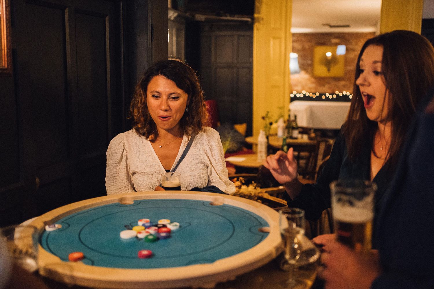 Two people playing carrom board game at night with one person reacting excitedly during gameplay.