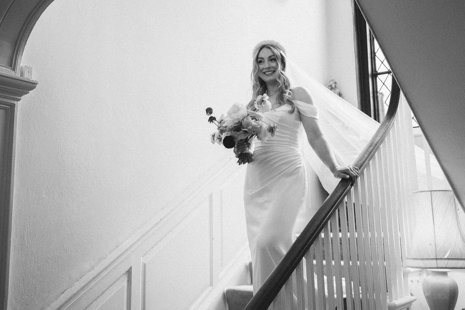 A bride in an elegant white gown descends a classic staircase while holding a bouquet in black and white photography.