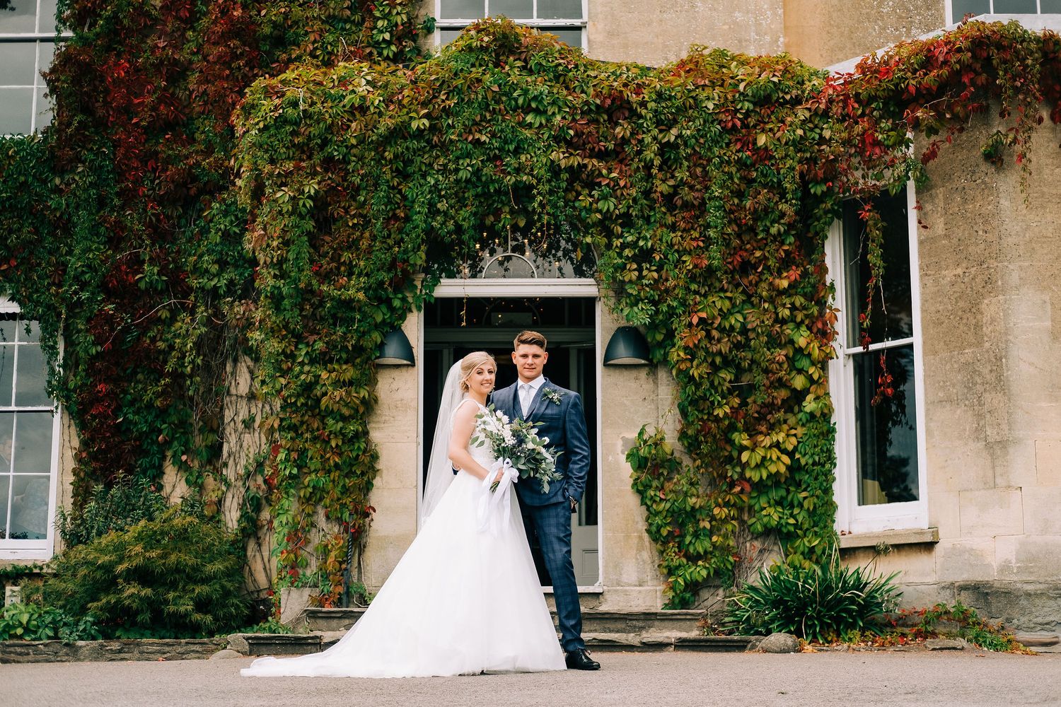 Wedding couple walks along ivy-covered building exterior.