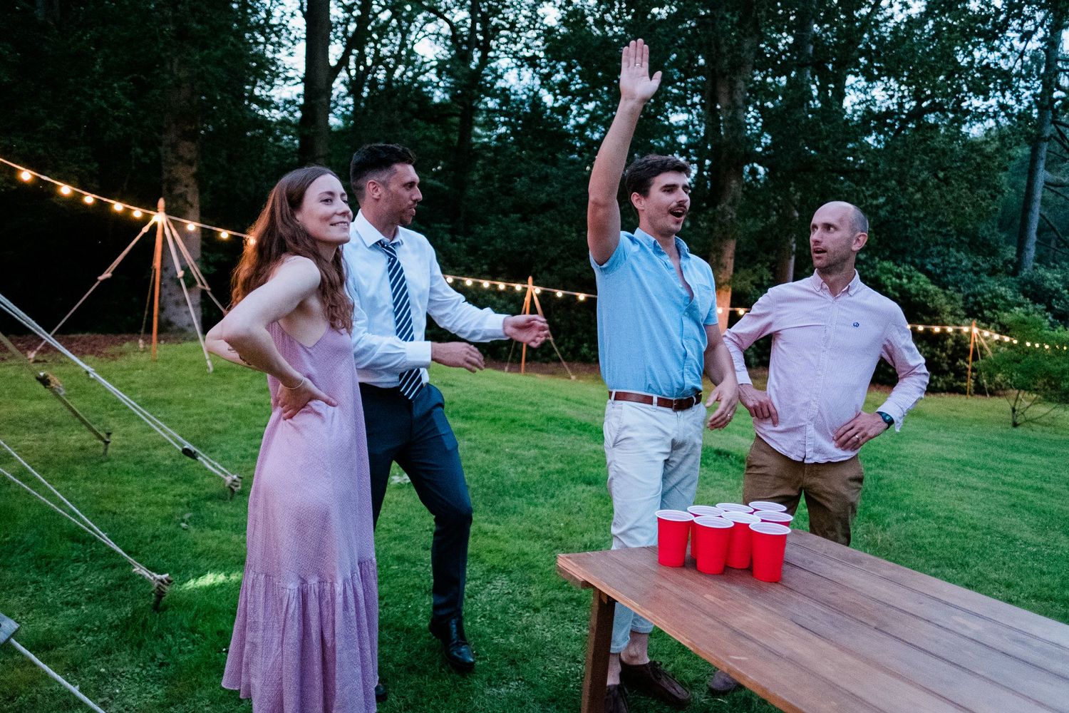 Party guests play outdoor lawn games and socialize on a grassy area near string lights.