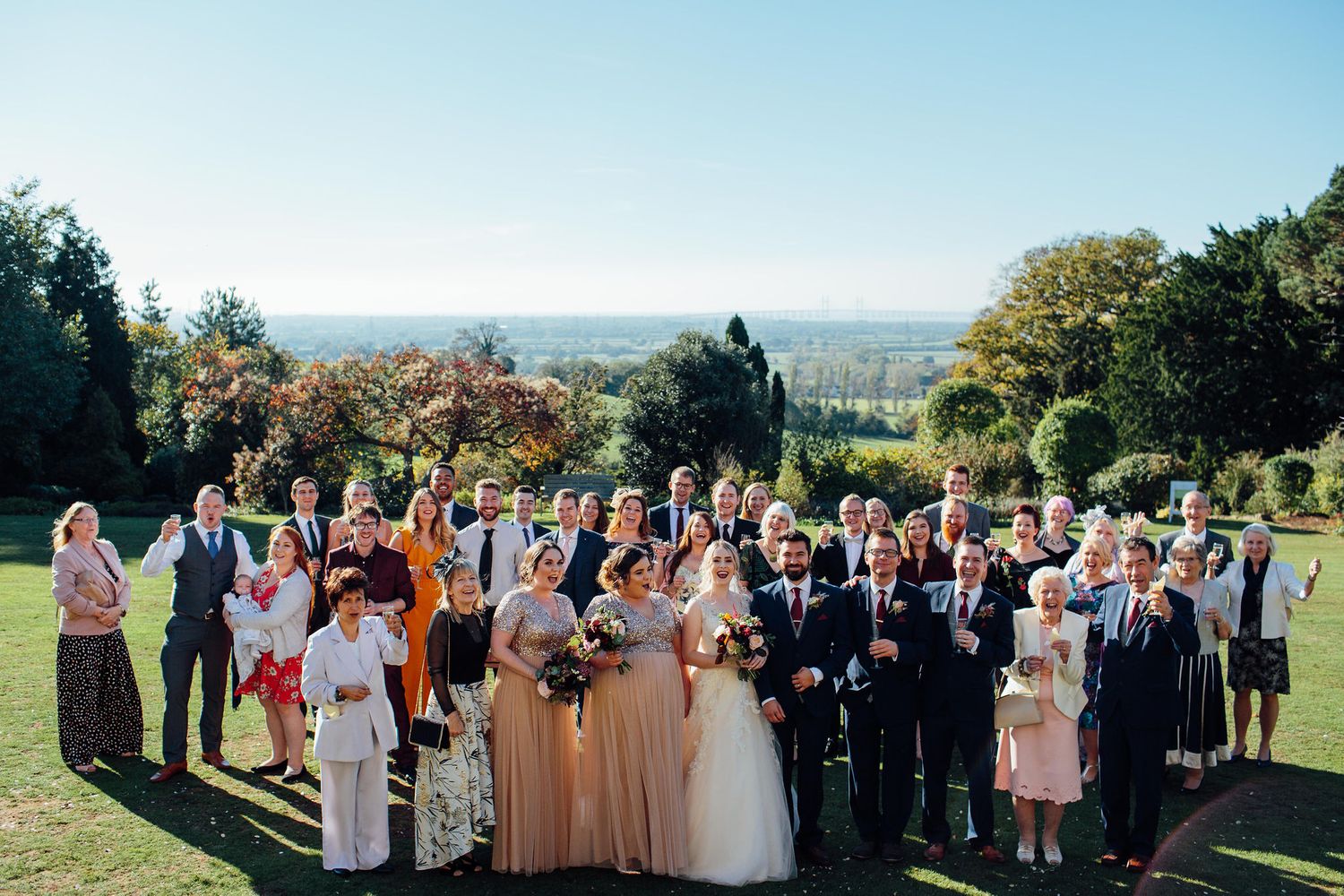 Wedding party and guests gather outdoors on a sunny day with rolling hills in the background.