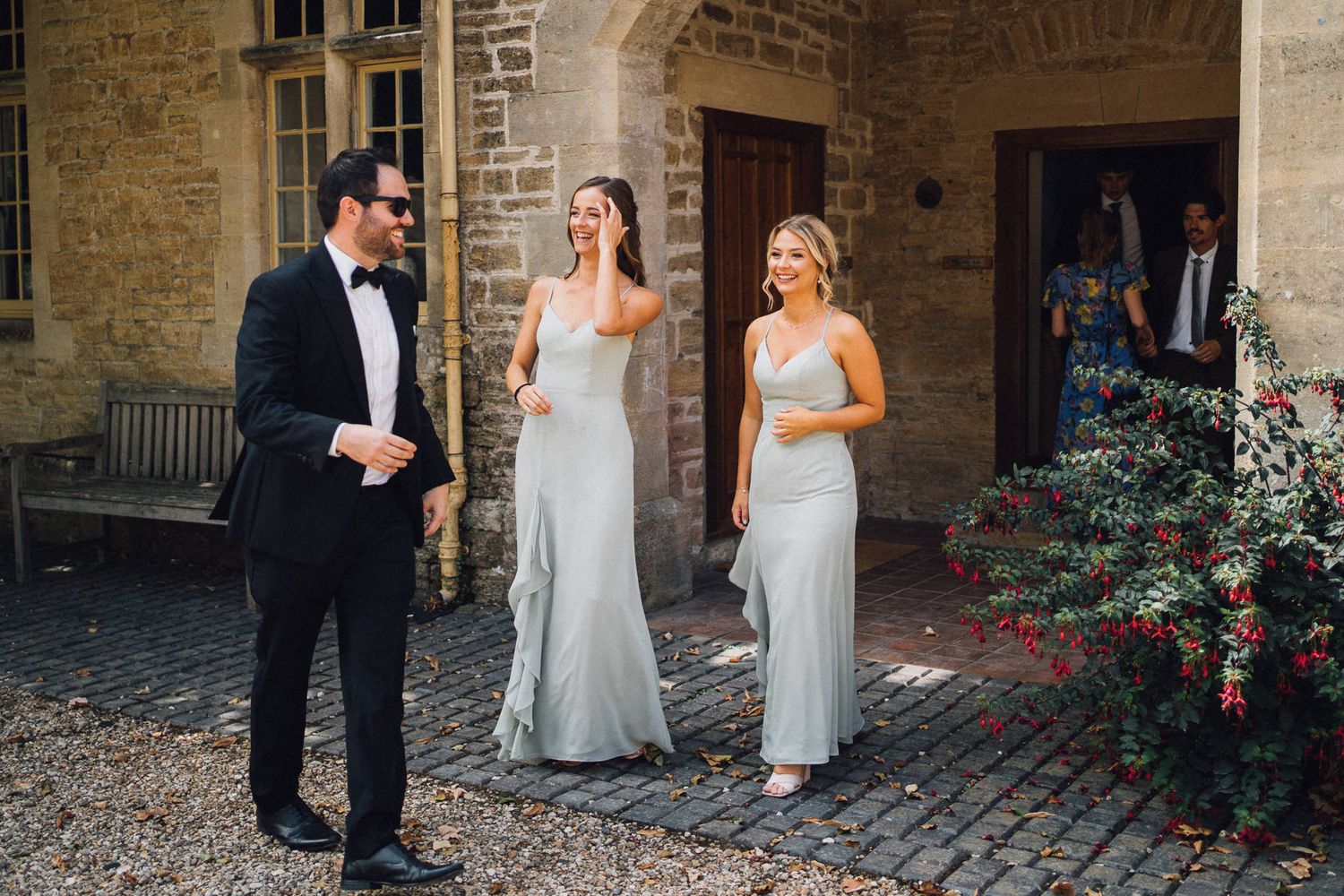 Wedding party members in sage dresses and formal attire gathered at stone venue entrance.