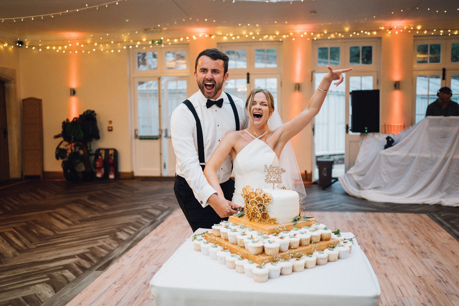 Newlyweds celebrate while cutting their tiered wedding cake surrounded by cupcakes.