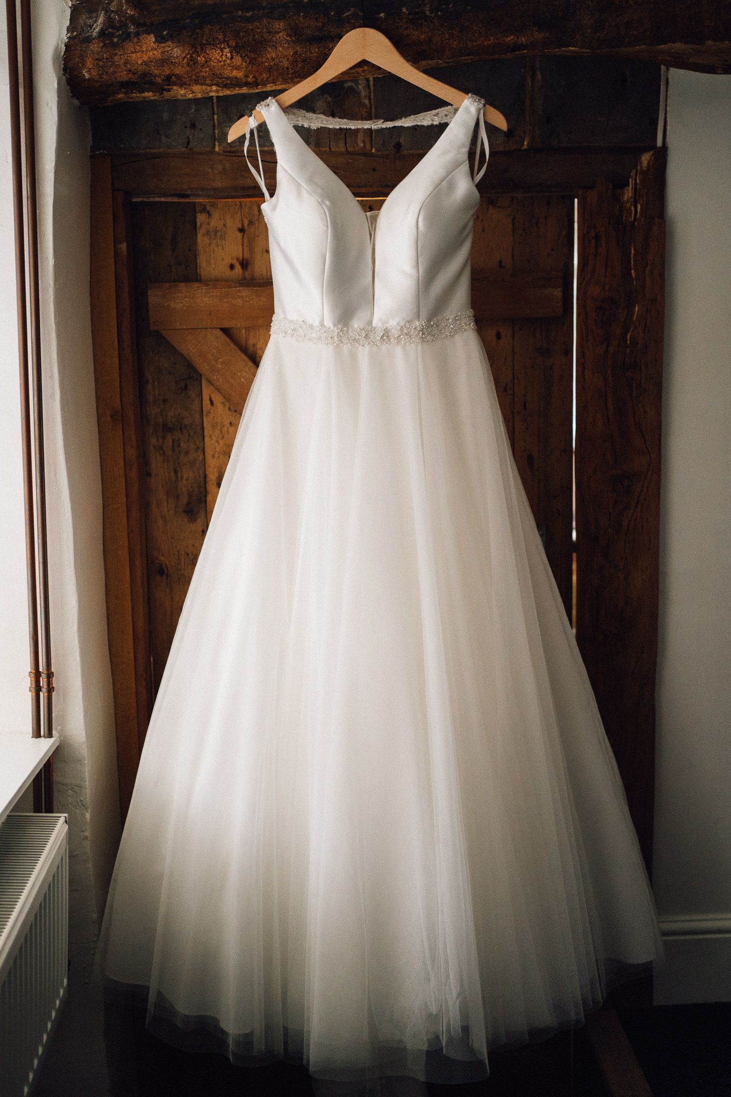 A white tulle wedding dress hangs elegantly on a wooden hanger against a rustic backdrop.