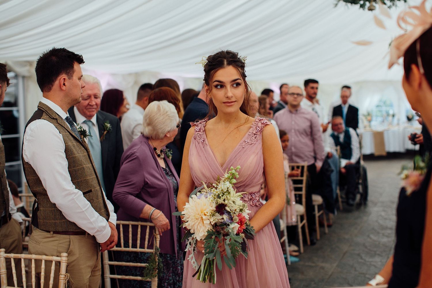 Wedding guests gather in a draped tent venue with wooden chairs and floral decorations.
