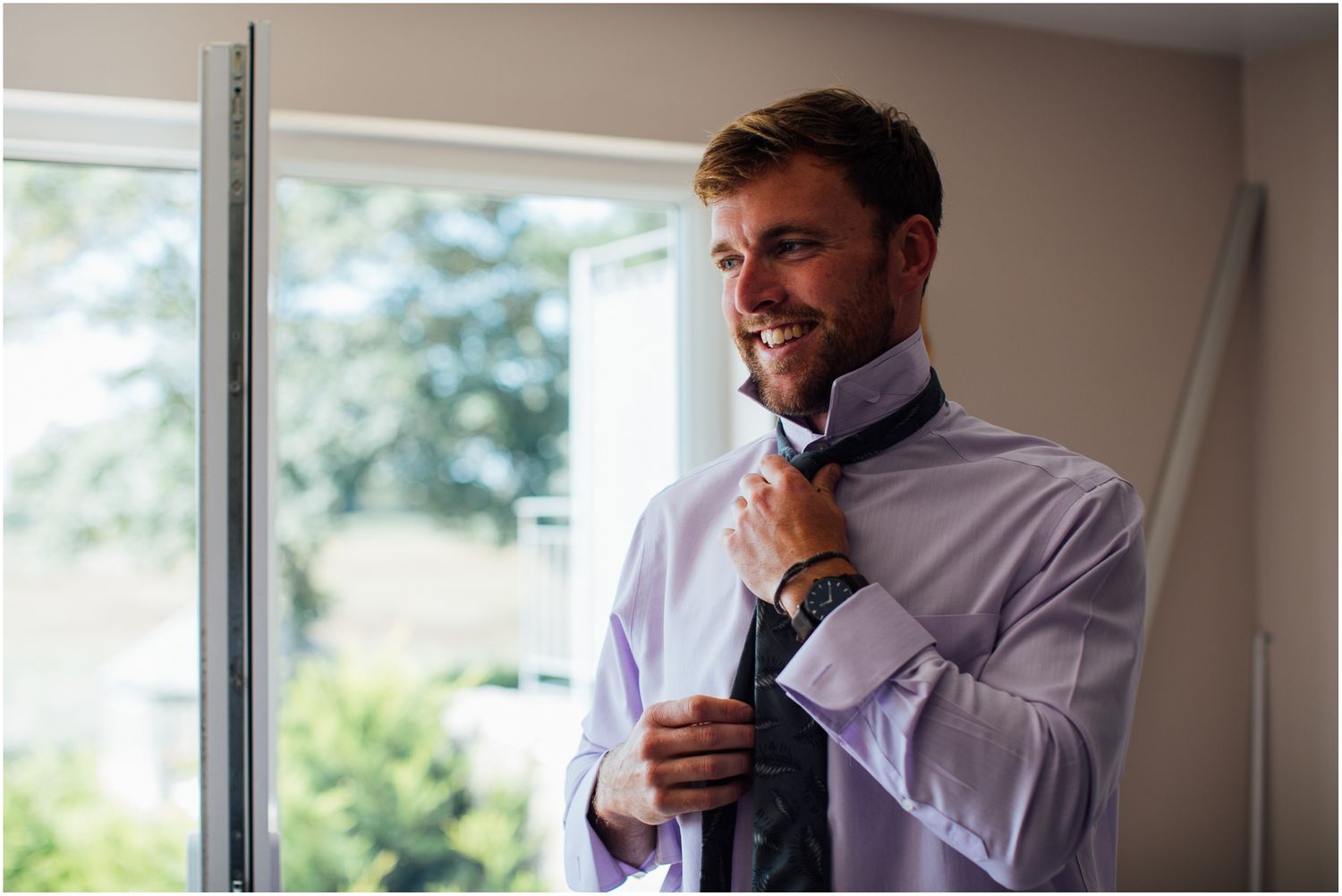A person adjusts their tie while getting dressed near a bright window.
