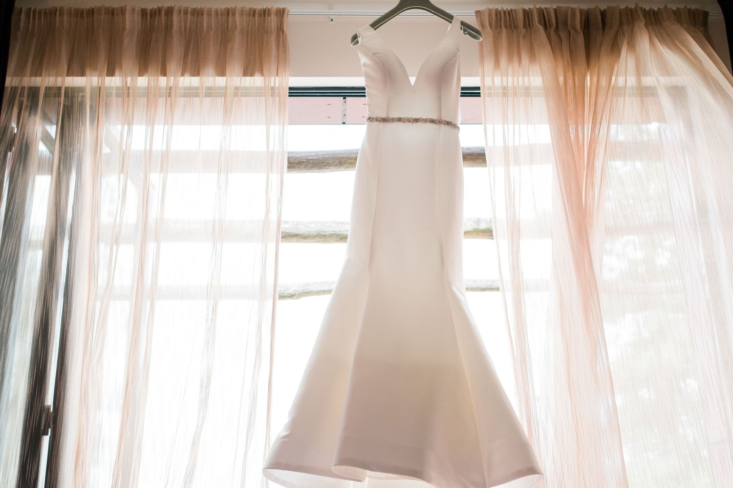 Wedding gown displayed in natural window light with flowing sheer curtains.