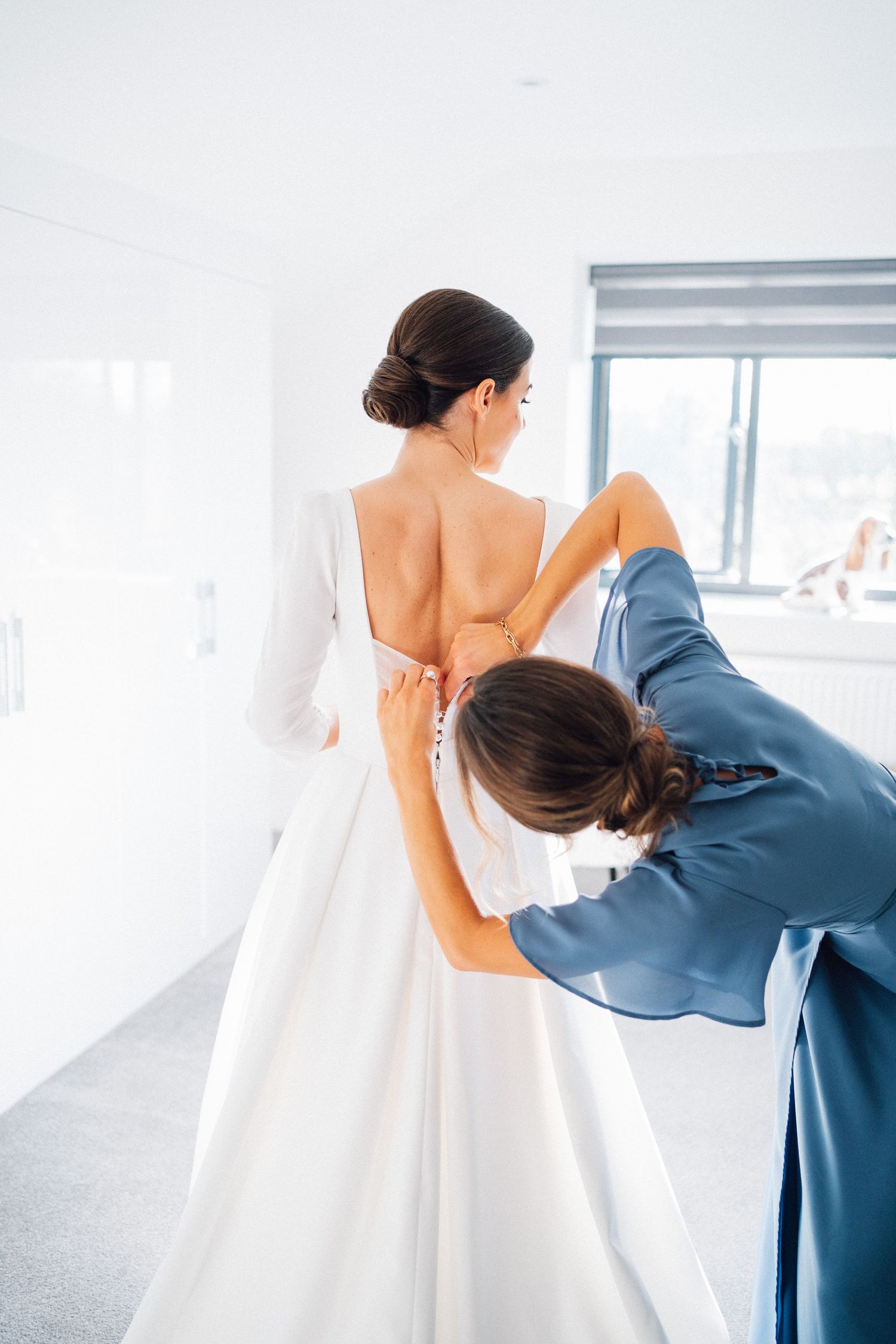 Wedding dress preparation scene with bride and helper adjusting elegant white gown in bright sunlit room.