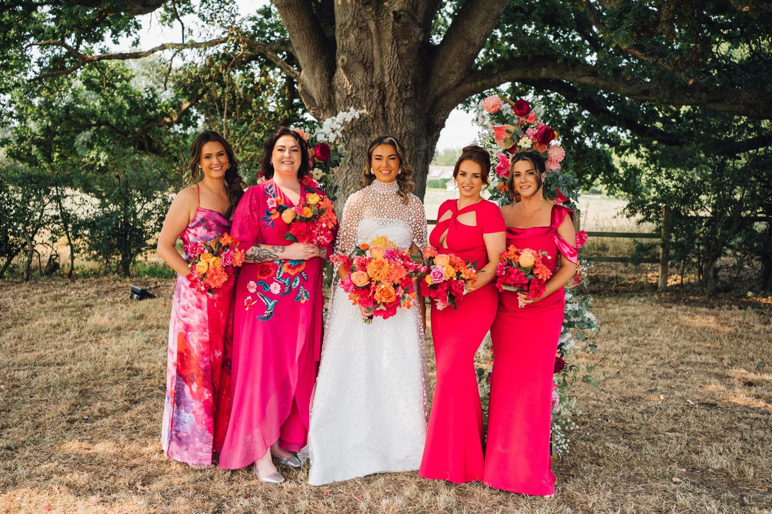A wedding party in bright pink bridesmaid dresses and white gown poses together outdoors under trees.