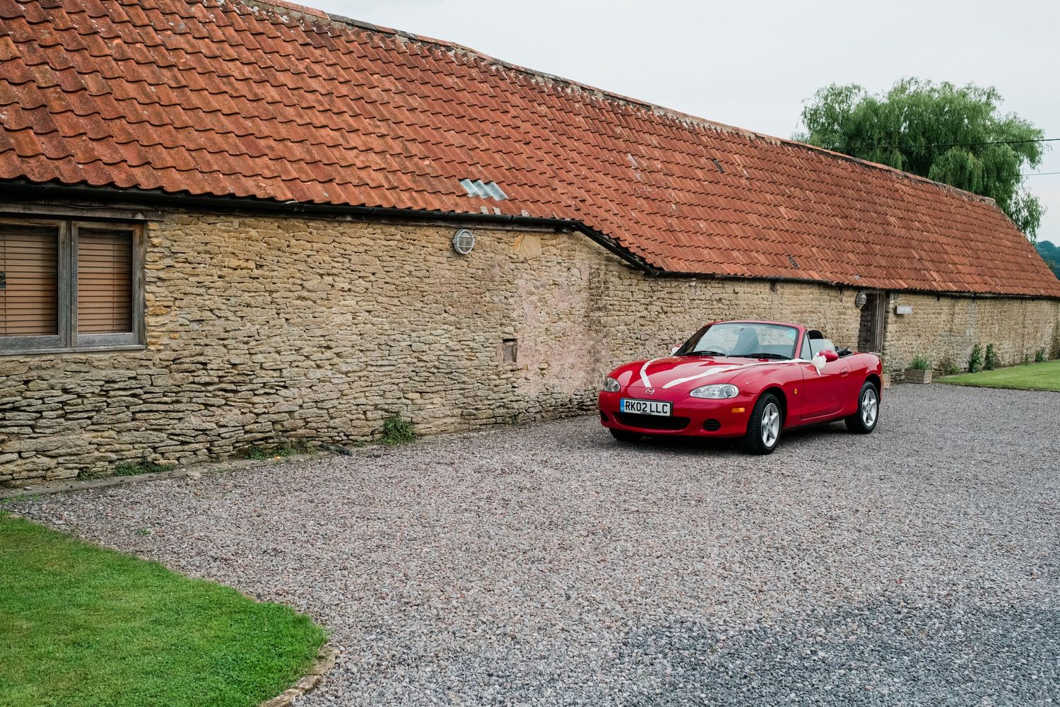 Red vintage sports car parked on gravel driveway alongside stone buildings with terracotta roofs.