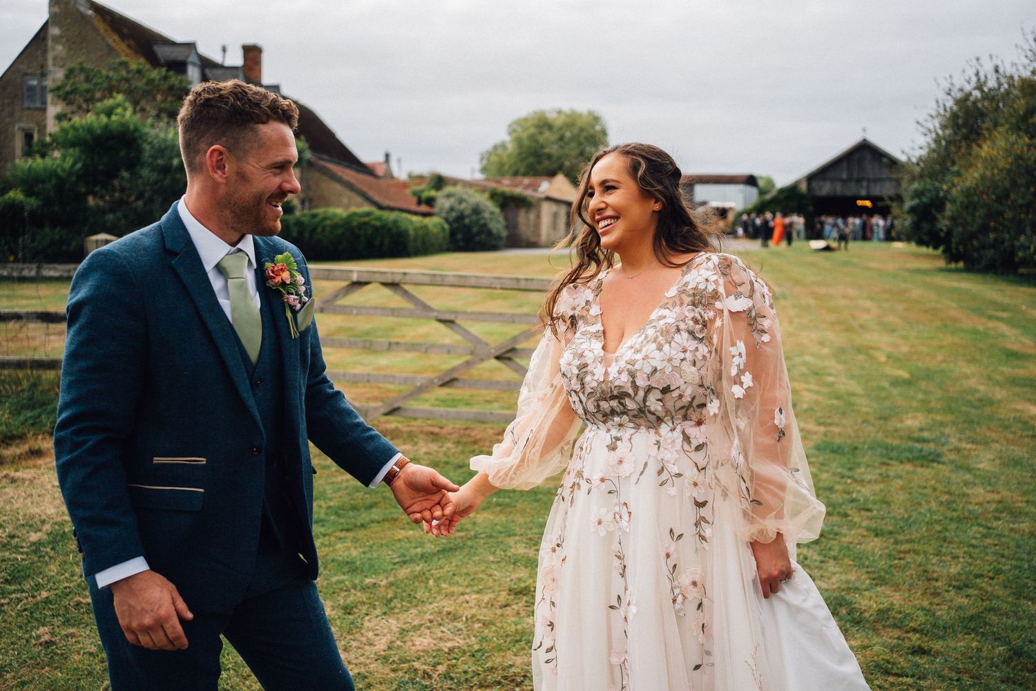 A couple celebrates their wedding day walking together on grassy grounds near farm buildings.