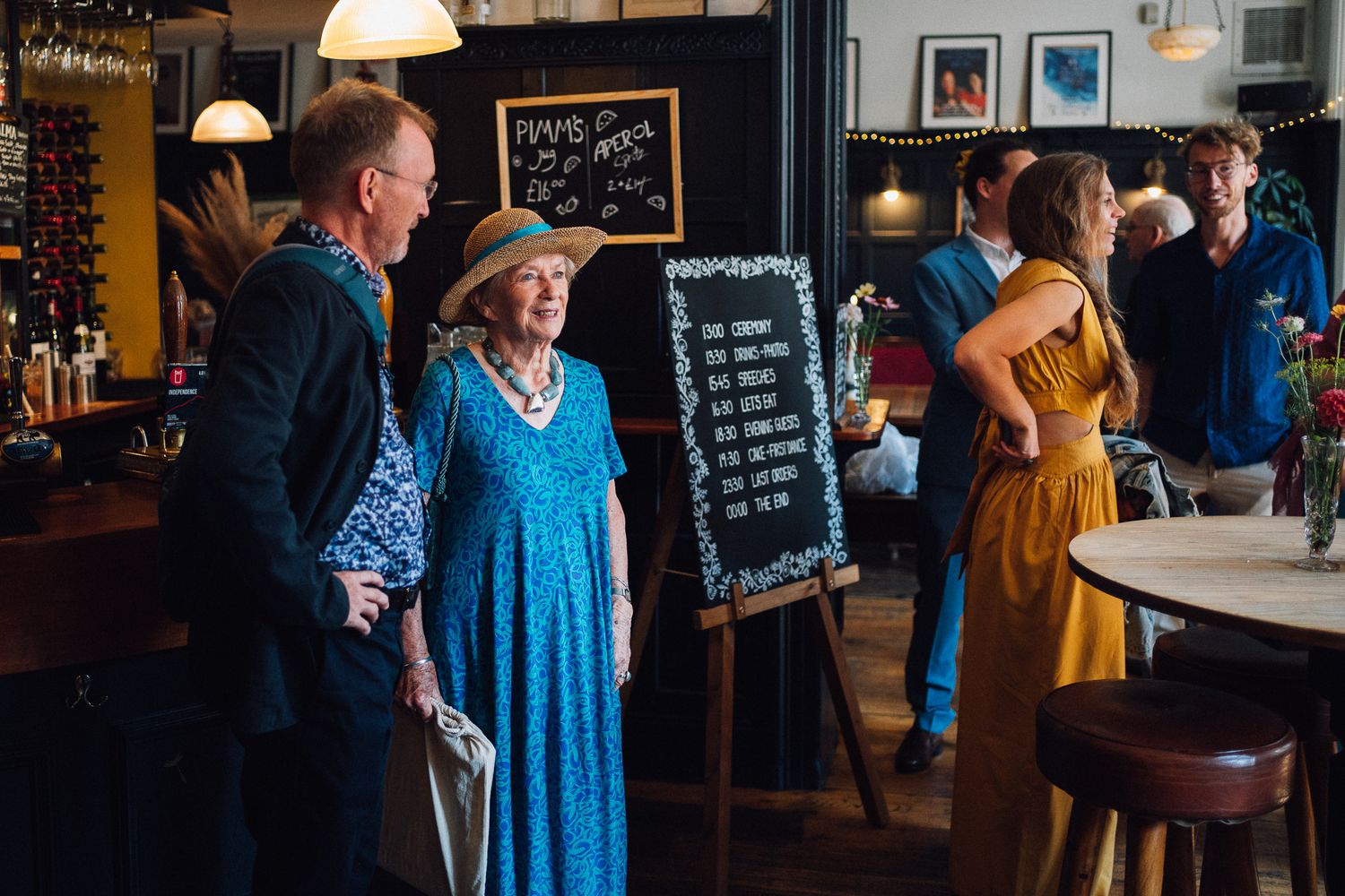 People mingle and socialize at a casual indoor event with chalkboard menus and pendant lighting visible in the background.