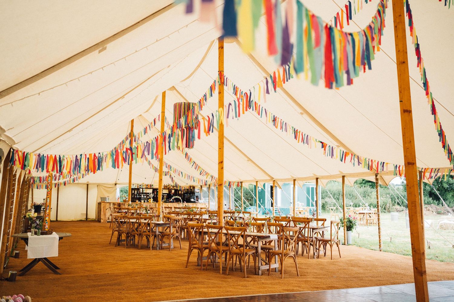 Colorful bunting flags decorate the ceiling of an elegant white wedding tent with wooden chairs and tables arranged below.