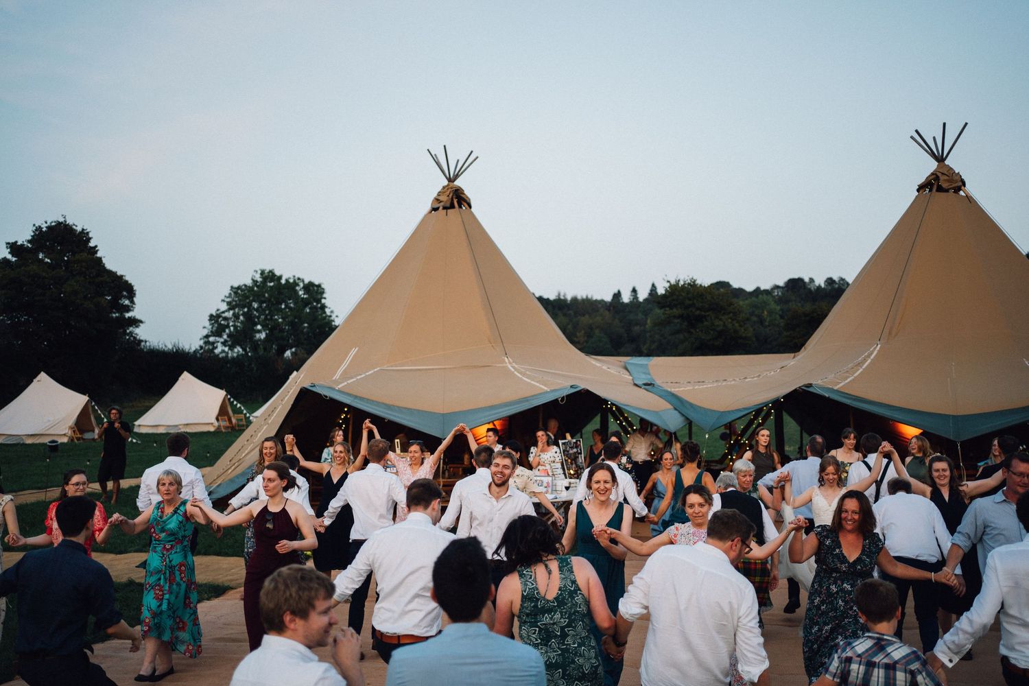 Guests dance and celebrate at an outdoor wedding reception with multiple tipi-style tents in the background.