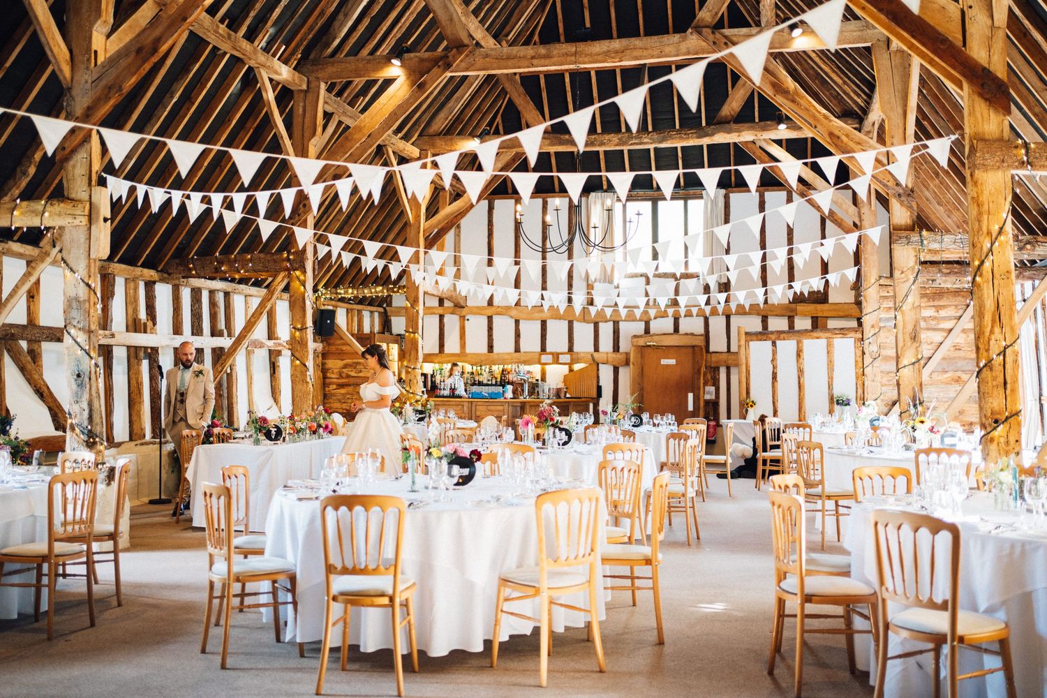 Long banquet tables set for a wedding reception inside a rustic barn with white bunting and wooden beams.