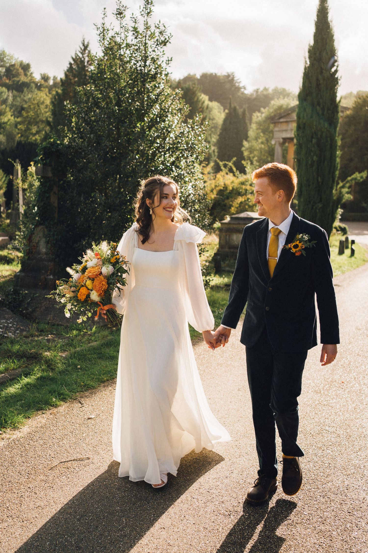 A couple walks along a sunlit garden path with cypress trees and greenery in the background.