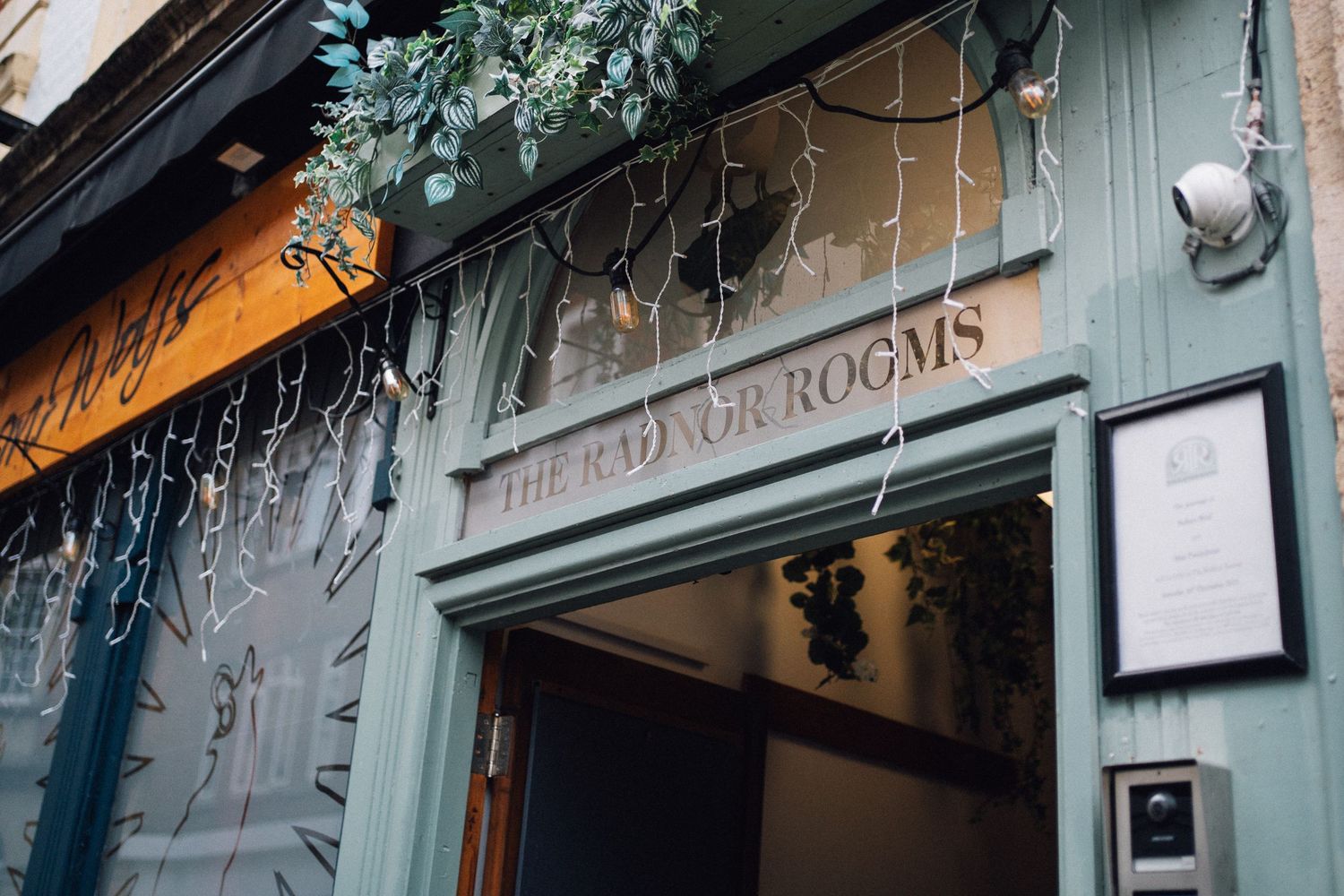 Sage green storefront entrance with hanging plants and a menu board displays 'The Good Year Rooms' signage.