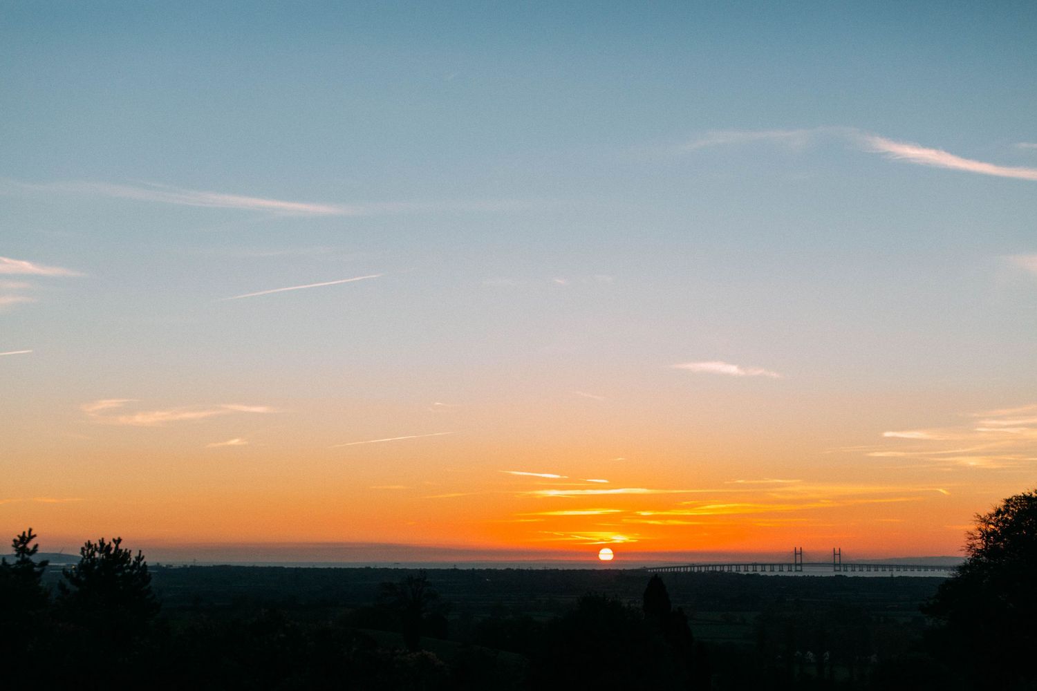 A vibrant orange and purple sunset over a dark silhouetted horizon with wispy clouds in the sky.