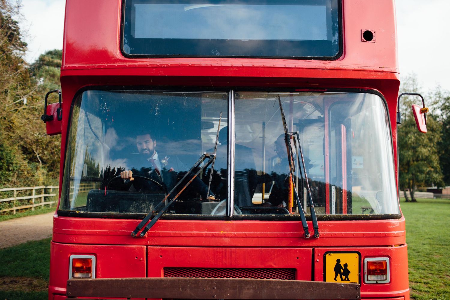 A red double decker bus is parked on a road with trees and grass visible in the background.