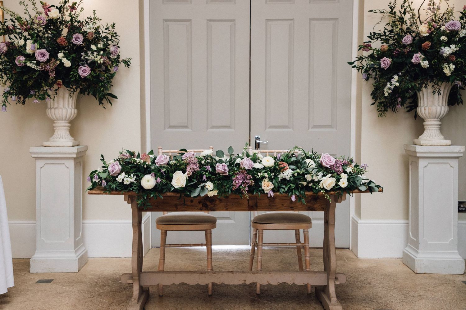 Rustic wooden tables adorned with pink roses and greenery arrangements against white paneled wall create elegant decor.