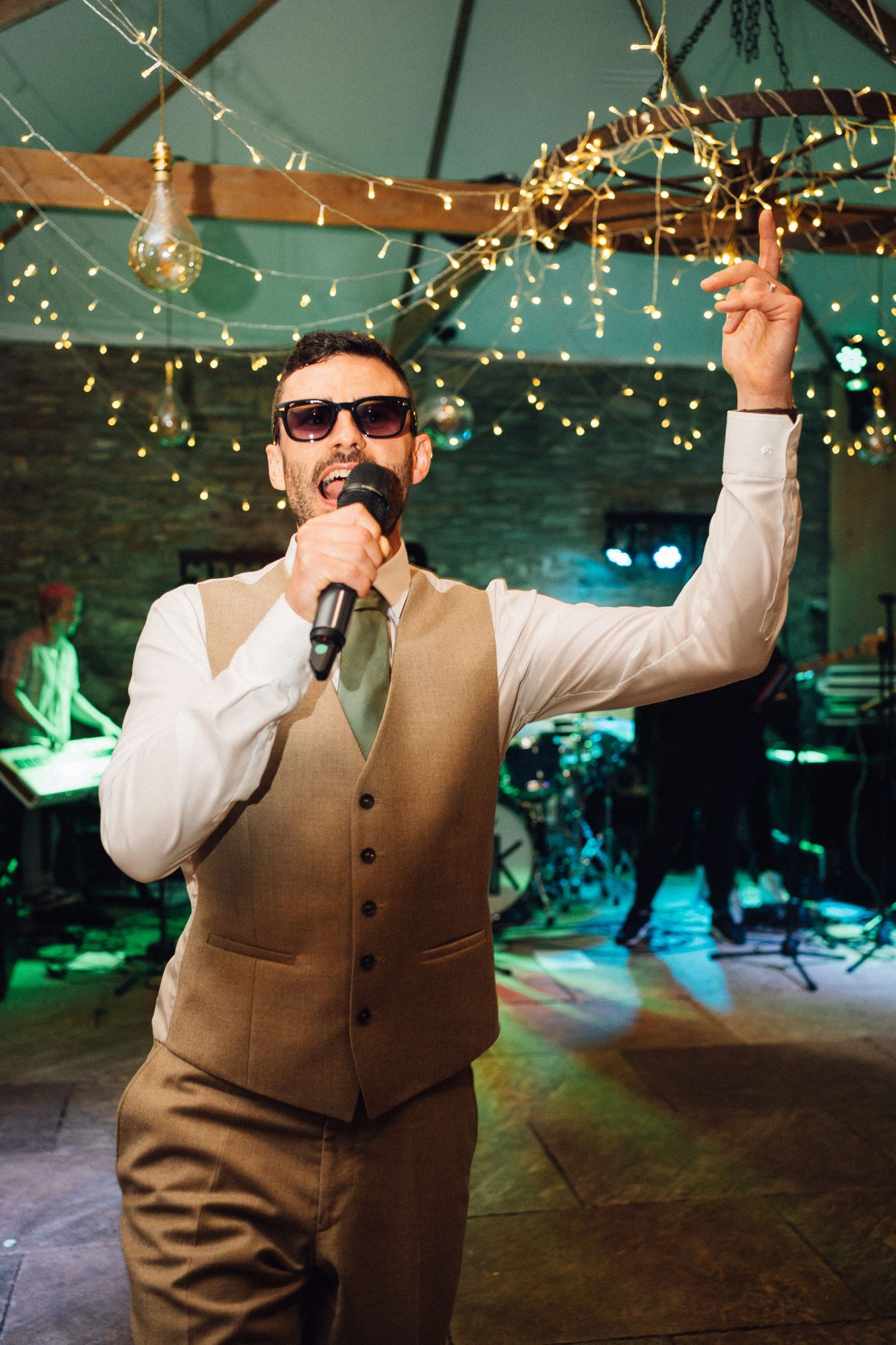 A performer in a brown vest and sunglasses singing under string lights at an indoor venue.
