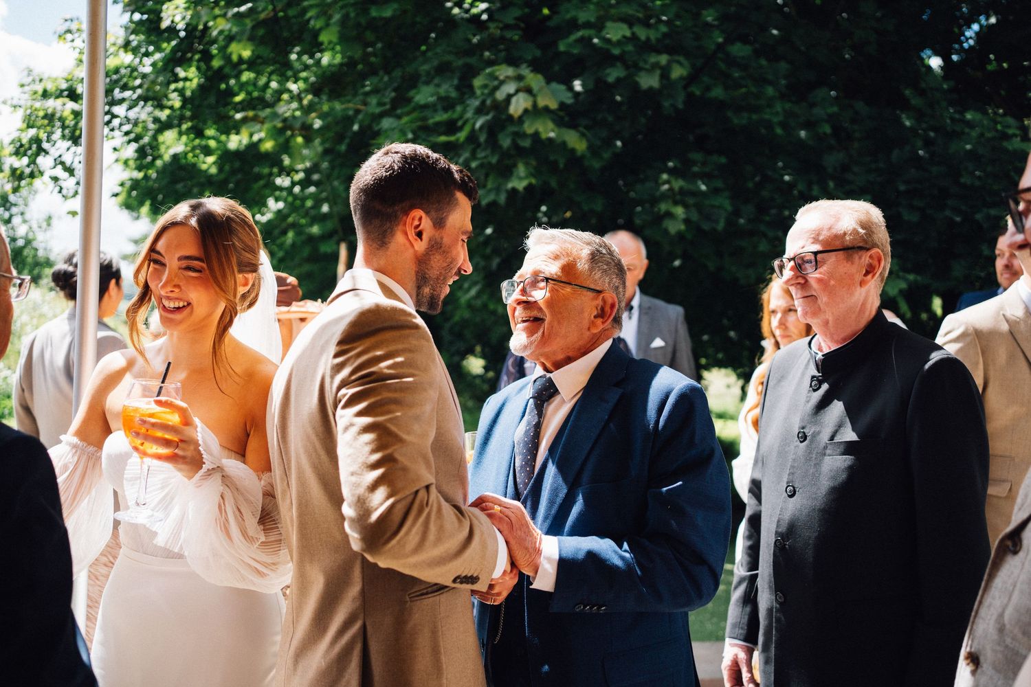 Wedding guests share joyful laughter during an outdoor celebration beneath green trees.