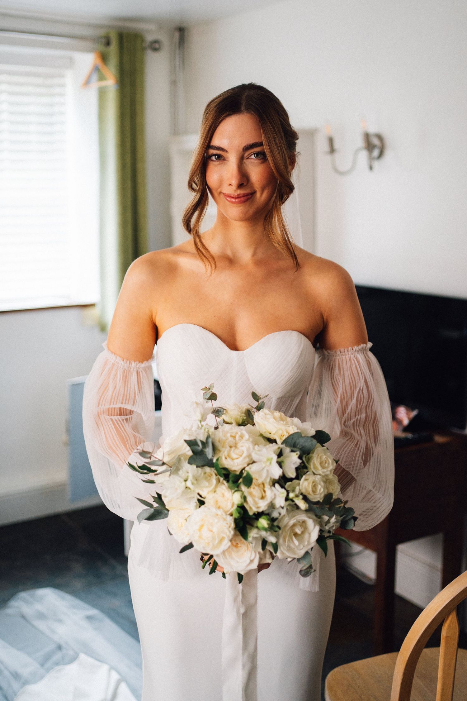 A bride in an elegant white off-shoulder wedding dress holds a lush bouquet of white flowers and greenery indoors.
