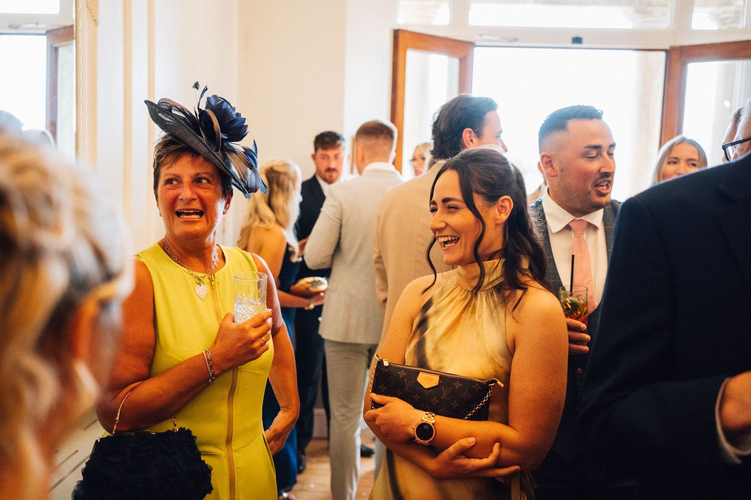 Wedding guests in formal attire mingling and socializing at an indoor reception venue.