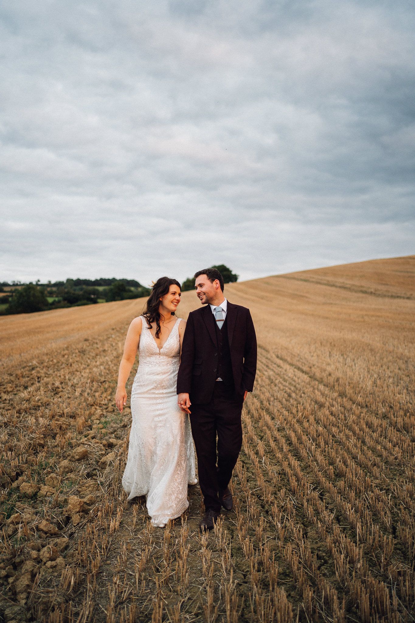 A couple in wedding attire walk together through a harvested wheat field under cloudy skies.