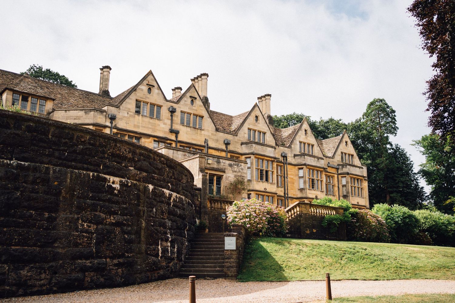 Grand Tudor-style stone mansion with multiple gabled roofs sits atop a grassy hill behind curved stone wall.