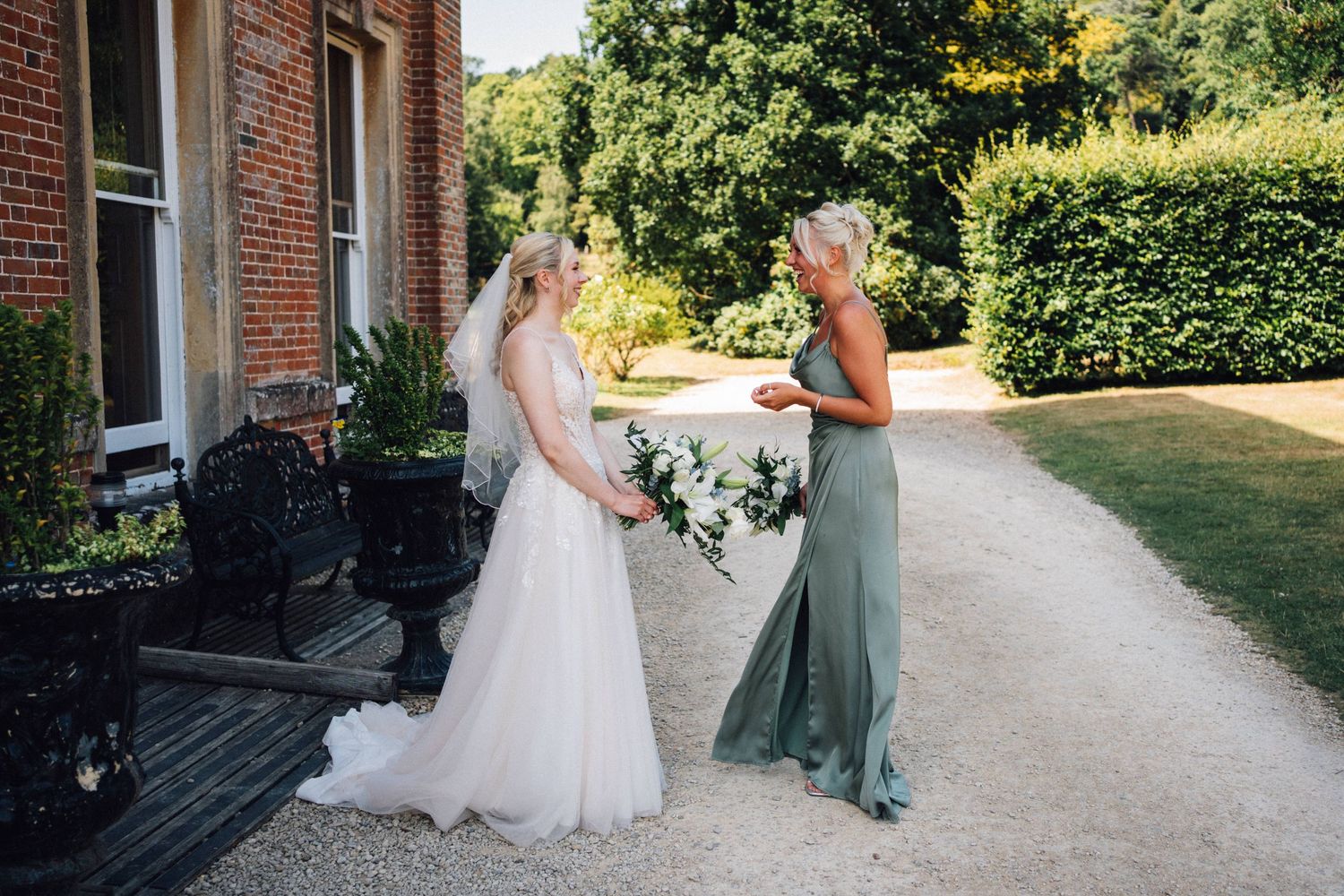 Wedding couple walks along a sunlit garden path bordered by brick buildings and lush greenery.