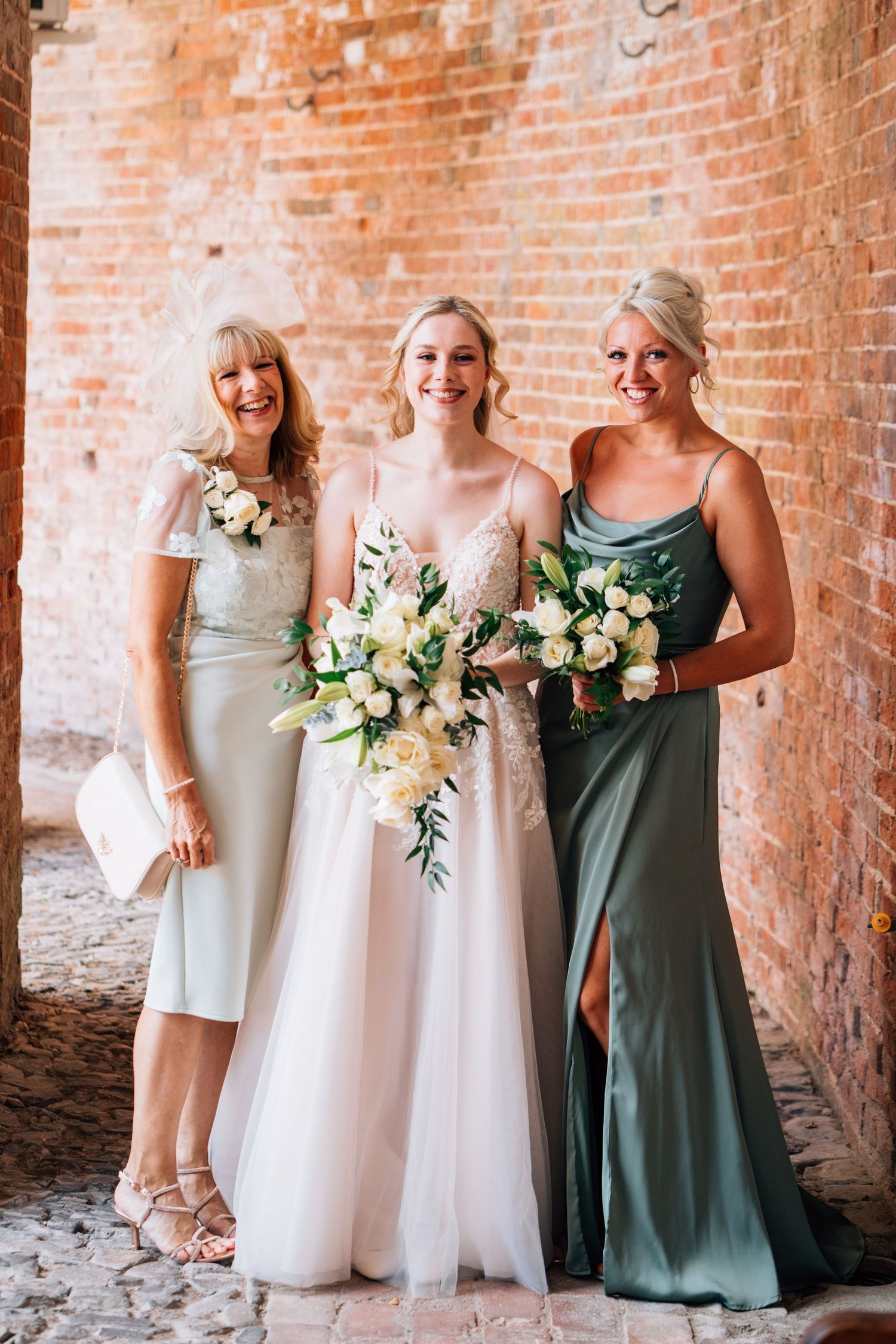 Wedding party in sage green and ivory dresses posing against exposed brick wall.
