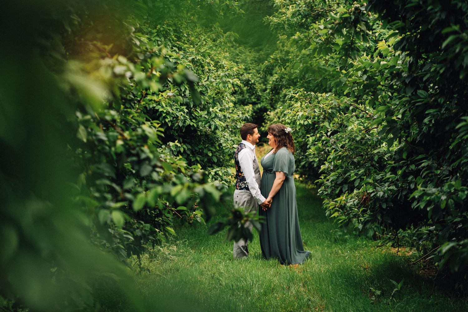 A couple shares a romantic moment in a lush garden setting surrounded by green foliage and natural light.