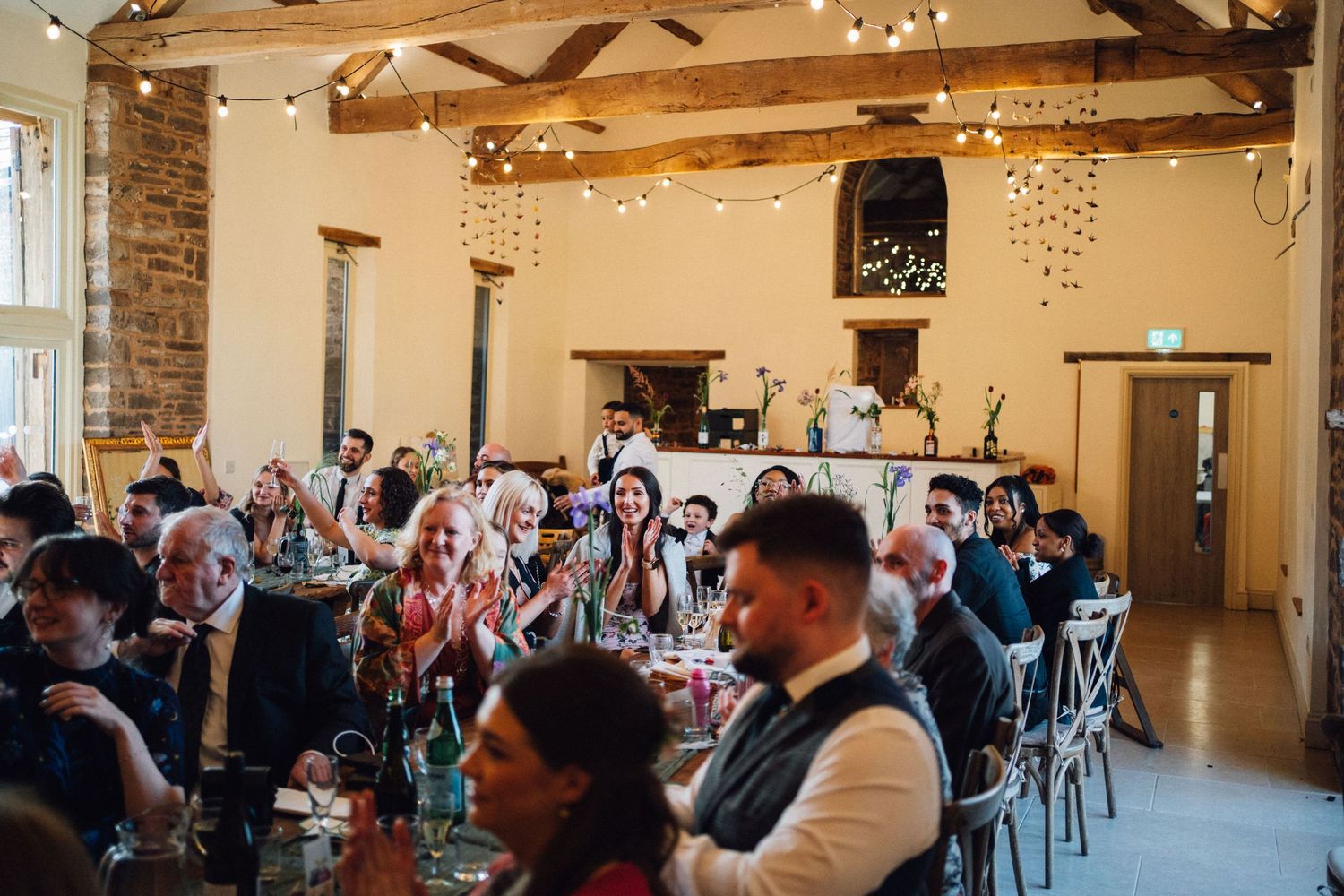 Wedding reception guests seated at long tables in a rustic barn venue with exposed wooden beams and fairy lights.