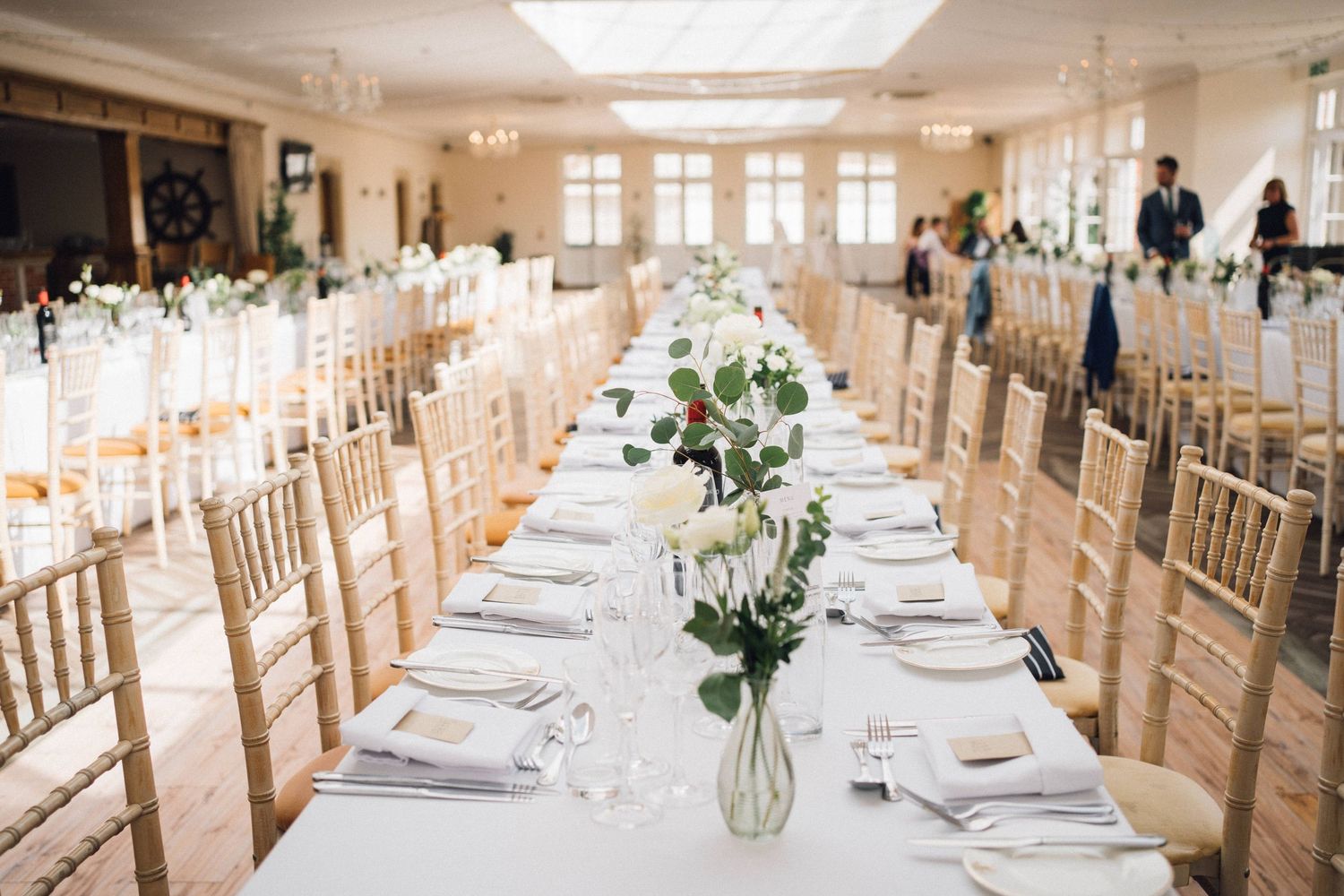 Elegant wedding reception venue with long banquet tables set with white linens and floral centerpieces in a bright room.