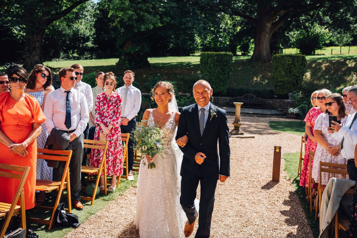 Wedding guests walk along a gravel path through a garden during an outdoor summer celebration.