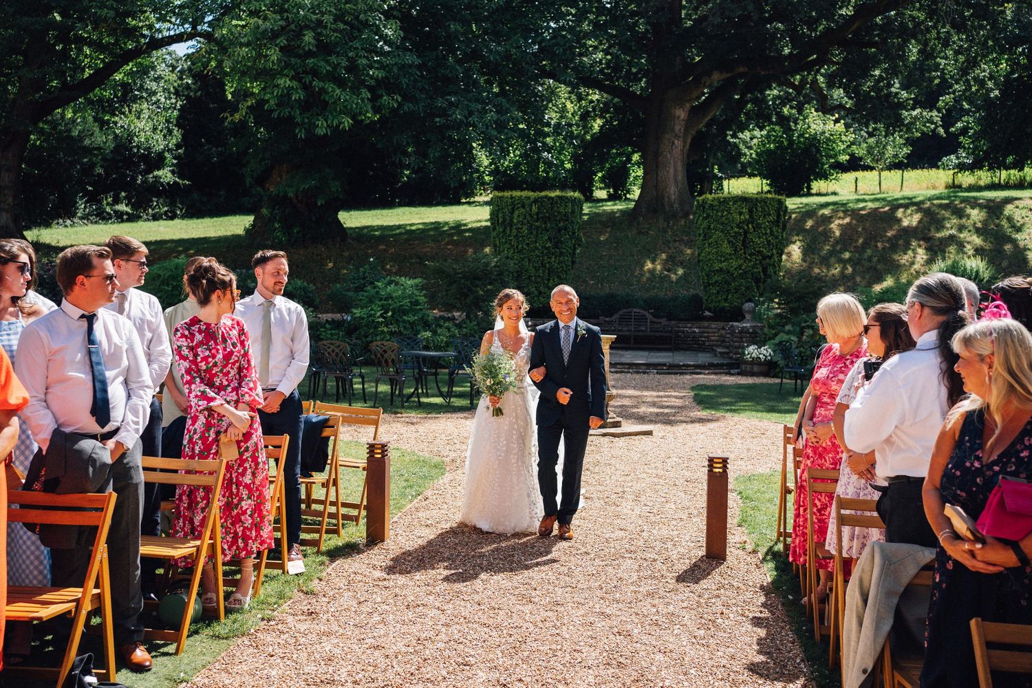 Wedding guests gather outdoors on a sunny day along a gravel path surrounded by lush greenery and trees.