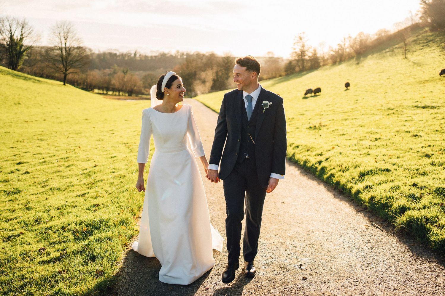 A couple walks hand in hand along a scenic countryside path with rolling green hills and grazing horses in the background.