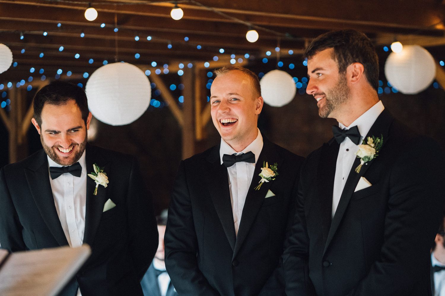 Wedding party members share laughs in a barn venue decorated with string lights and white paper lanterns.