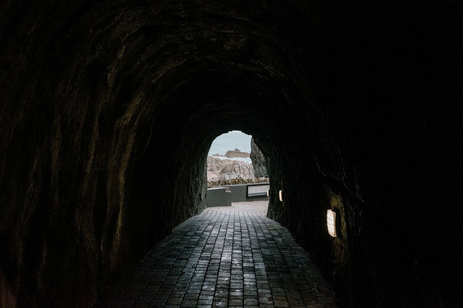 Dark stone tunnel with cobblestone floor leading to daylight at the end.
