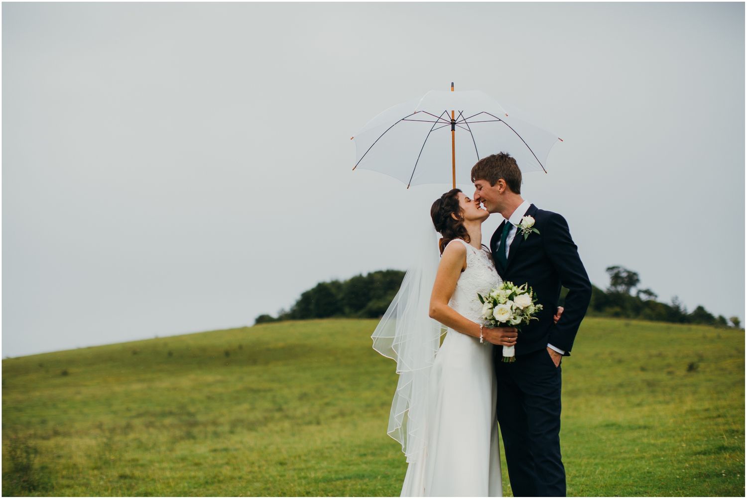 Wedding couple shares romantic moment under clear umbrella in overcast weather.