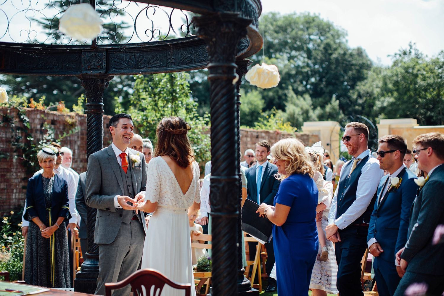 Wedding guests seated in rows at an outdoor ceremony with brick walls and greenery in the background.