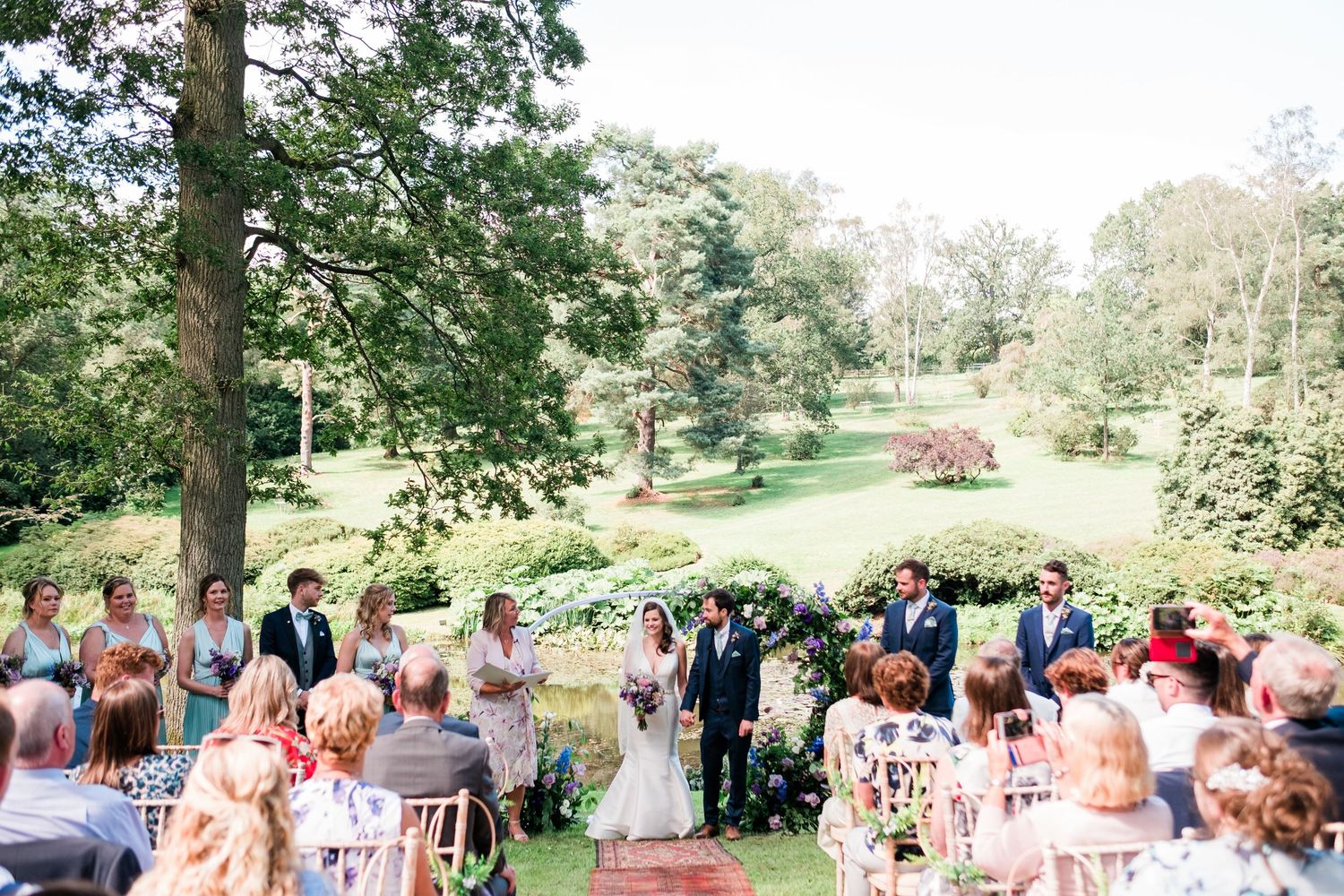 Outdoor wedding ceremony setup with white chairs on a lush green lawn surrounded by trees.
