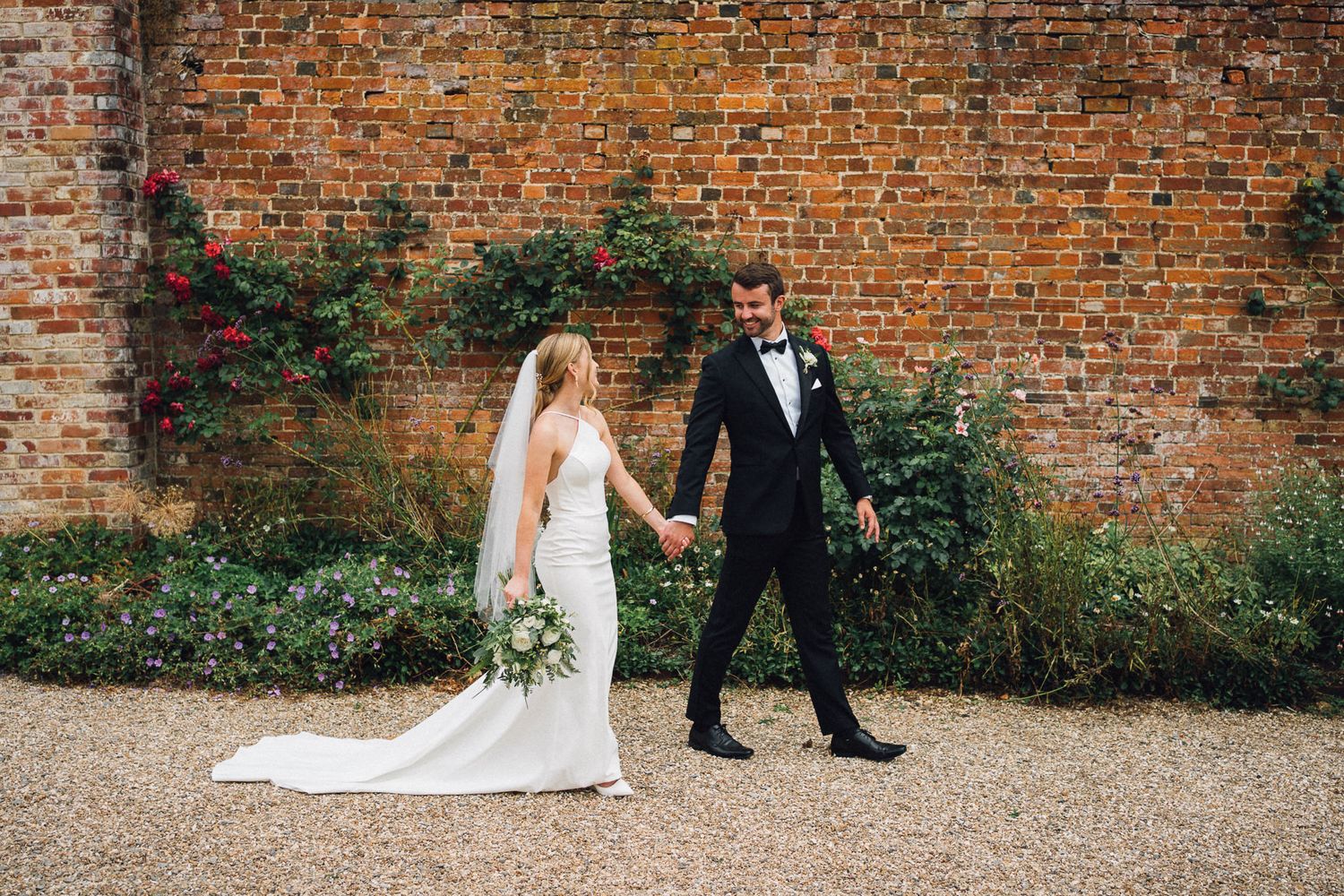 Couple walking along rustic brick wall during outdoor wedding photoshoot.