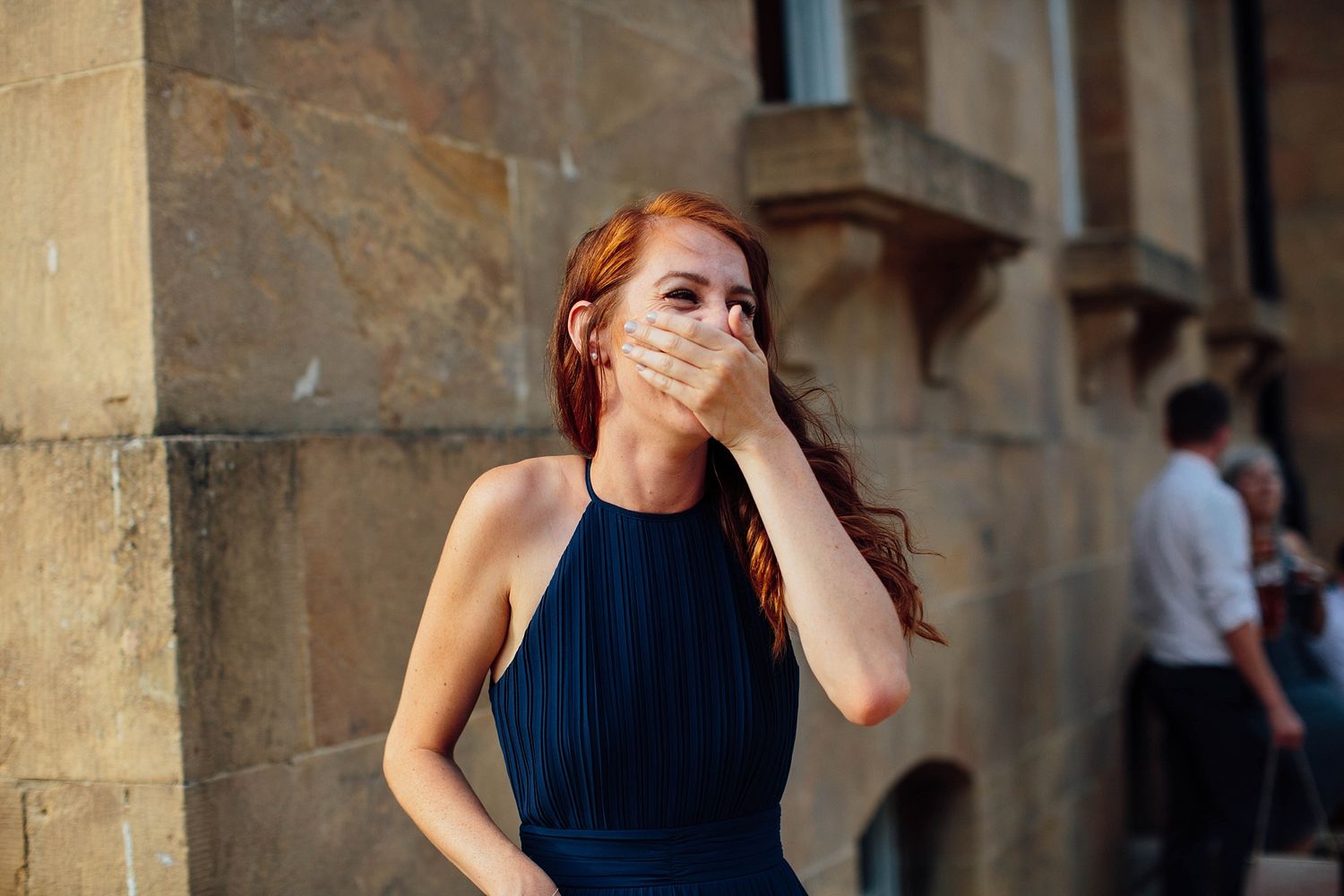 A woman in a navy halter dress shows emotion outside a historic stone building.