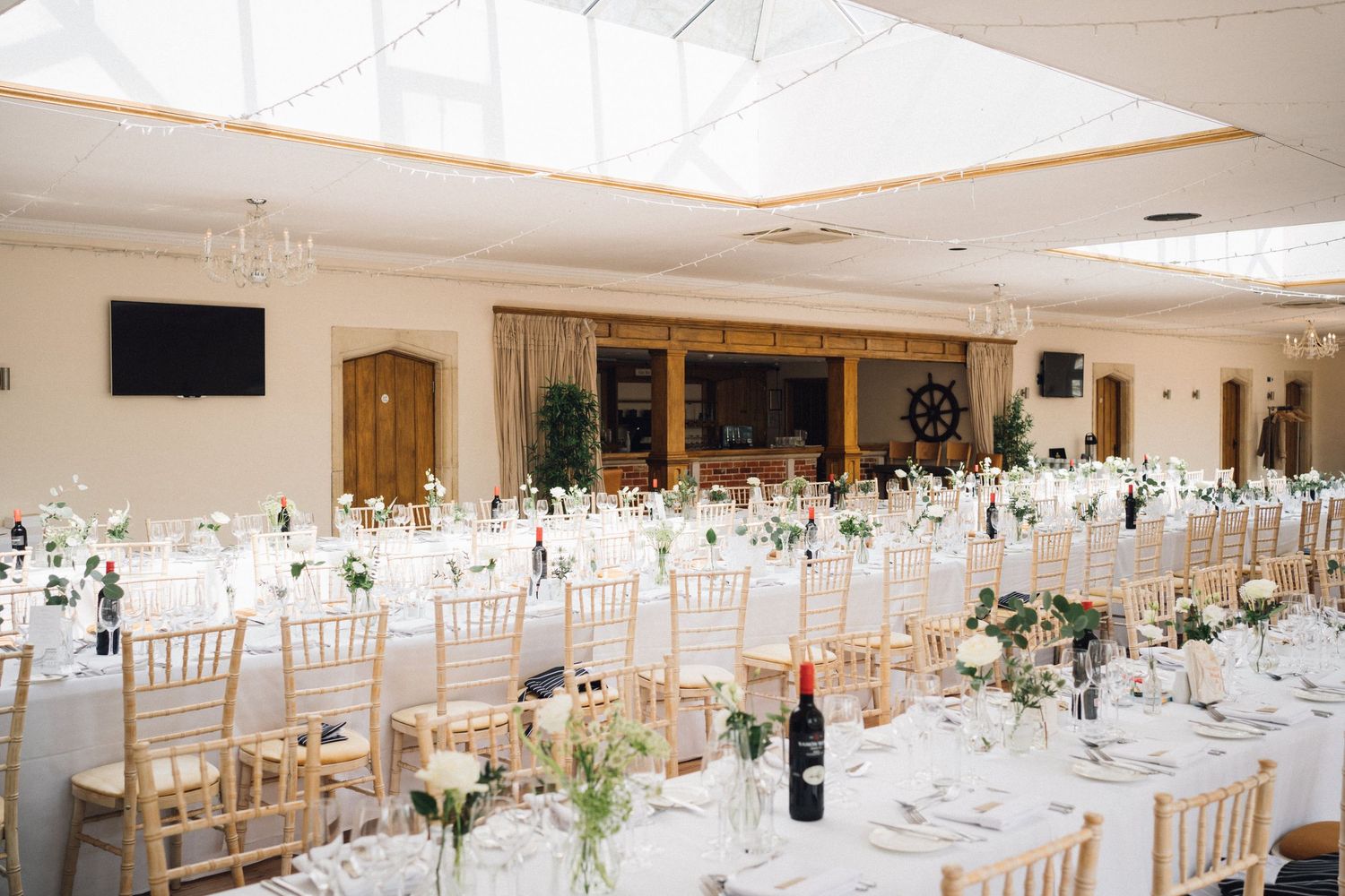 Elegant wedding reception hall with long white tables, gold chiavari chairs, floral centerpieces, and a skylight ceiling.