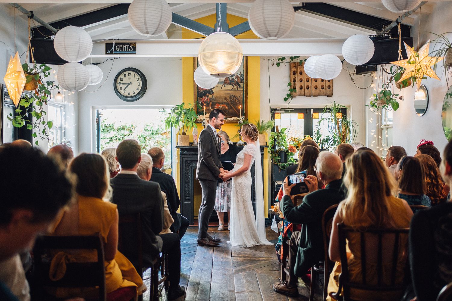 Couple exchanges vows at indoor wedding ceremony with white lanterns and guests seated in a decorated venue.