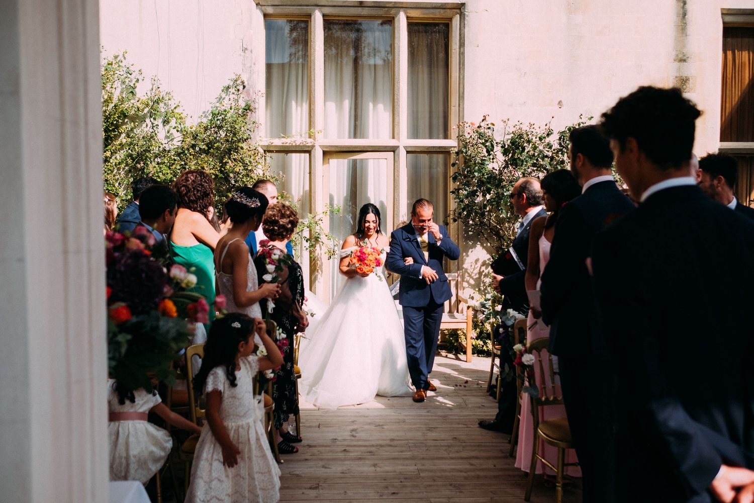 A wedding ceremony processional taking place in a bright, flower-decorated corridor.