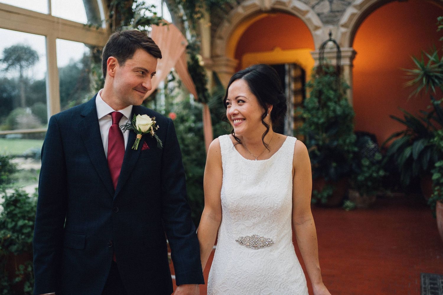 Wedding couple embraces in front of stone archways and orange walls with potted plants.